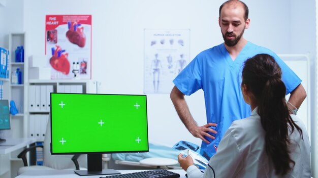 Assistant giving doctor x-ray image while working on computer with green screen monitor in hospital cabinet. Desktop with replaceable screen in medical clinic while doctor is checking patient radiogra