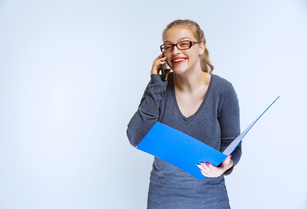 Assistant talking to the phone white checking the blue folder and taking notes, and looks very happy.