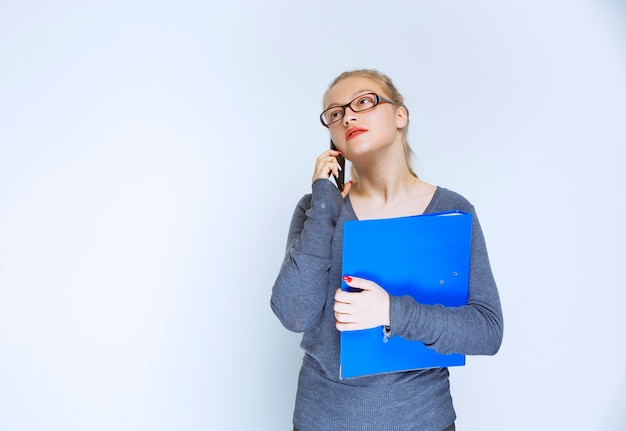 Assistant with eyeglasses holding a blue folder and talking to the phone.