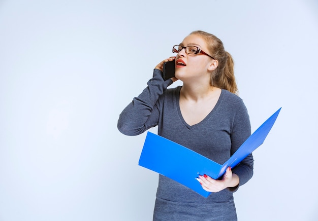 Assistant with eyeglasses holding a blue folder and talking to the phone.