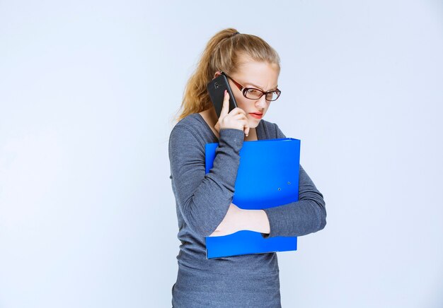 Assistant with eyeglasses holding a blue folder and talking to the phone.