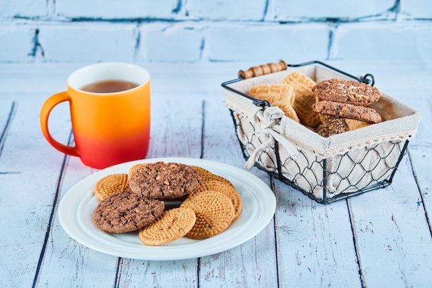 Assorted biscuits in plate and basket and a cup on blue table.