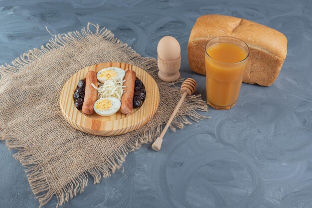 Ассорти из блюд для завтрака рядом с буханкой хлеба, персиковым соком, вареным яйцом и ложкой меда на мраморном столе.