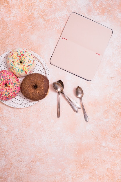 Assorted donuts with chocolate frosted; pink glazed and sprinkles donuts on white plate spoon and blank notebook against rustic backdrop