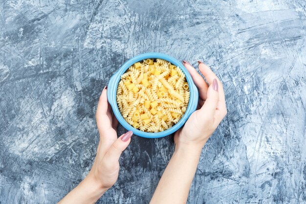 Assorted pasta in a bowl and hand holding a bowl of pasta top view on a grey plaster background