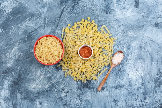Assorted pasta in a bowl with salt in wooden spoon, pepper flakes flat lay on a grungy plaster background