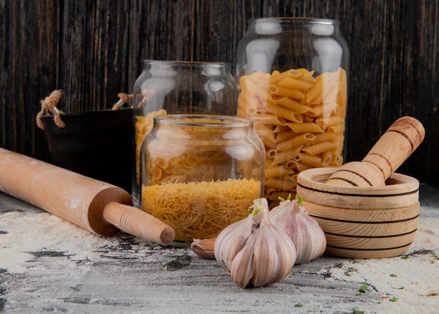 Assorted raw italian pasta in glass jars with garlic and rolling pin on the table with flour