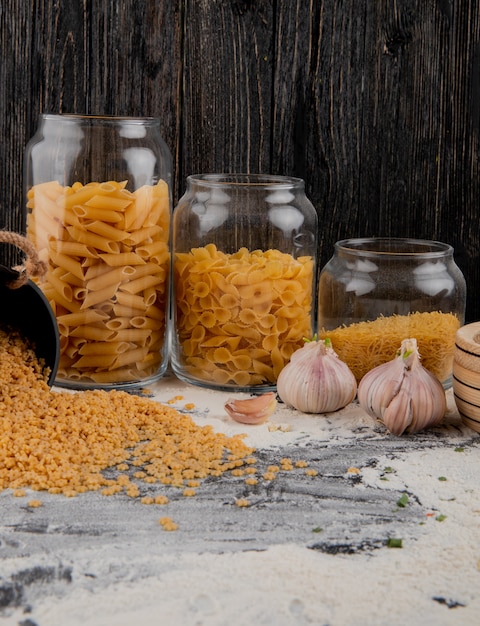 Assorted raw italian pasta in glass jars with garlic on the table with flour