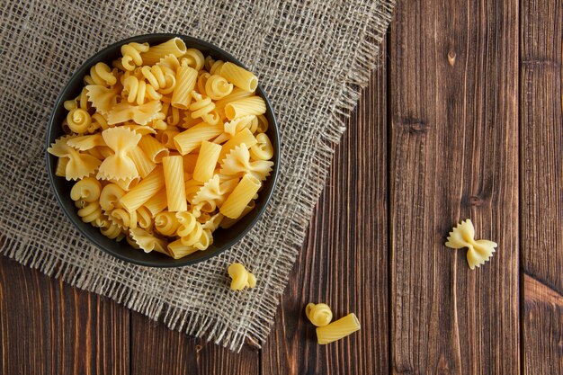 Assorted raw pasta in a bowl on wooden and piece of sack background. flat lay.