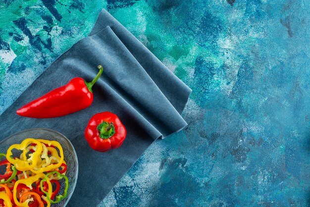 Ассорти из нарезанного перца в миске рядом с красным перцем на кусках ткани, на синем фоне.