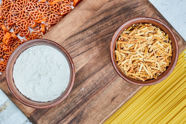 Assorted uncooked pastas and a bowl of flour on the wooden board.