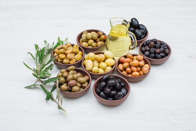 Assorted variety of olives in a clay bowls with olive leaves and a jar of olive oil high angle view on white wood