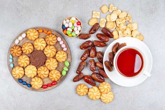 Ассортимент печенья, бисквитов, фиников и конфет с чашкой чая на мраморной поверхности.