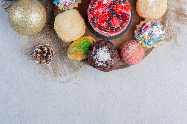 An assortment of desserts on a wooden board adorned with bauble and pine cone on marble surface