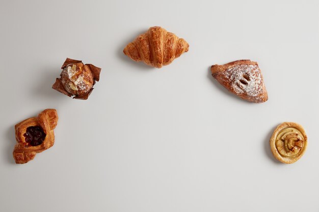 Assortment of freshly baked sweet bakery products. Buns, croissant, roll, muffin arranged in half circle against white background. Copy space in middle of shot. Puff pastry. Bakery foodstuff