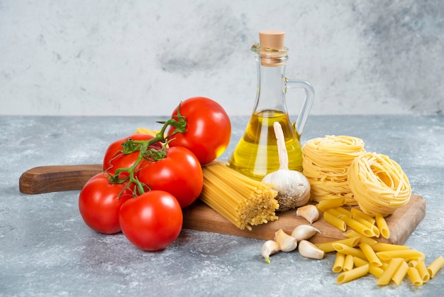 Assortment of raw pasta, olive oil and tomatoes on wooden board.