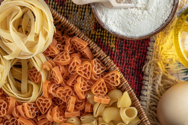 Assortment of raw pasta in wooden basket with flour and egg.