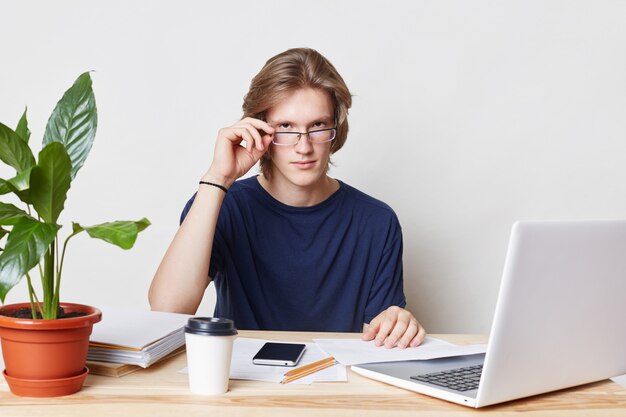 Assured male enterpreneur looks seriously through glasses, surrounded with modern laptop