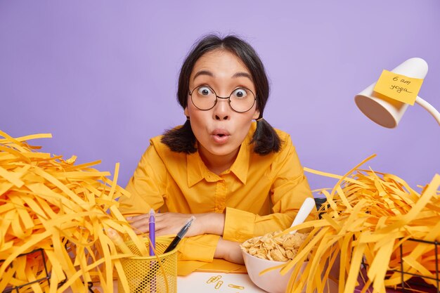 Astonished Asian schoolgirl stares shocked at front works all evening on project work surrounded by cut paper wears round spectacles yellow shirt sits at desktop isolated over purple wall