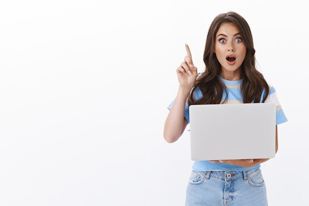 Astonished attractive modern female entrepreneur create awesome concept, hold laptop, raise index finger eureka gesture, gasping add suggestion, white wall