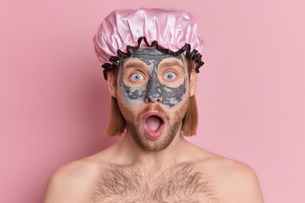 astonished bearded man applies clay mask on face stare with eyes popped out widely opened mouth wears protective bath hat.