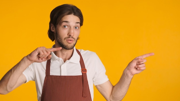 Astonished bearded man wearing apron pointing aside with fingers over colorful background Young waiter showing space for text
