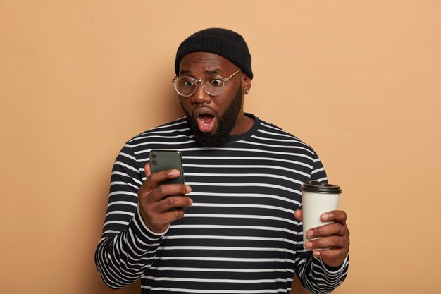 Astonished bearded young man stares at modern cellular, wears black hat and striped jumper