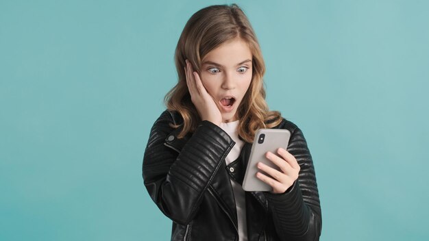 Astonished blond teenager girl getting message from friend on smartphone looking stunned over blue background