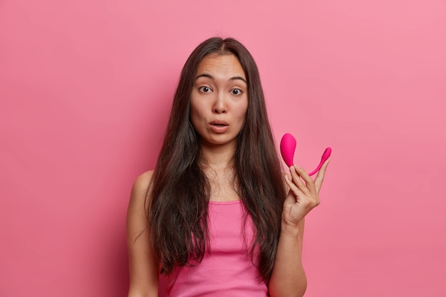 Astonished brunette female holds modern remote app controlled smart vibrator, visits sex shop and buys necessary accessories to satisfy herself, dressed casually, poses against pink wall