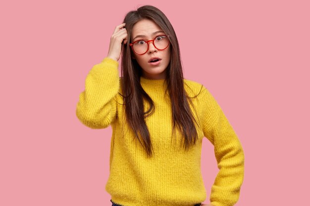 Astonished brunette woman scratches head, looks surprisingly, wears spectacles and yellow clothes