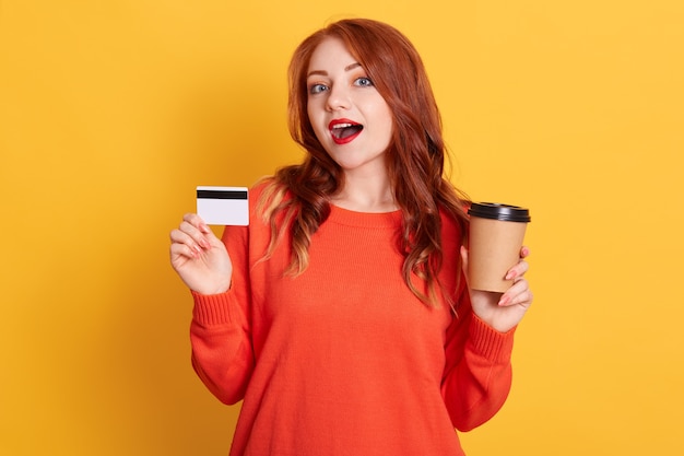 Astonished buyer finding offer online, holding take away coffee and credit card, has surprised facial expression, lady with red lips and wavy hair