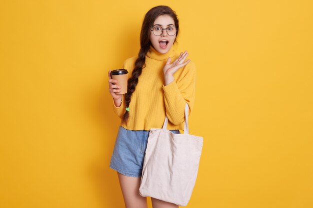 Astonished buyer with long pigtail holding takeaway coffee and shopping bag in hands