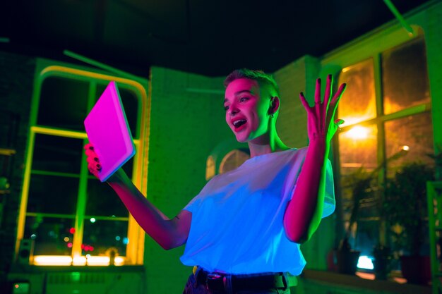 Astonished. Cinematic portrait of stylish woman in neon lighted interior. Toned like cinema effects, bright neoned colors. Caucasian model using tablet in colorful lights indoors. Youth culture.