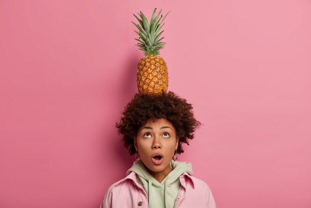 Astonished curly haired African American woman looks above, keeps mouth opened, carries pineapple on head, wonders how she can, wears hoodie and jacket, poses against pink pastel wall.