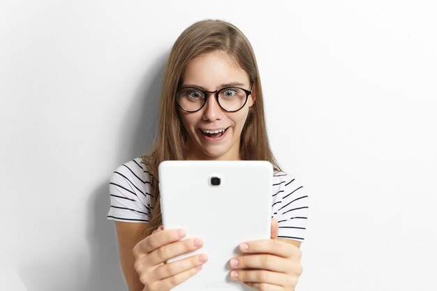 Astonished emotional funny teenage girl in stylish glasses holding generic digital tablet and opening mouth widely, being shocked while reading news or watching shocking content on internet