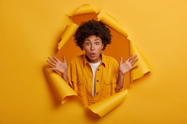 Free photo astonished fascinated ethnic girl raises palms, cannot believe in horrible situation happened with her, dressed in stylish clothing, poses at ripped paper hole, gets stunning chance.