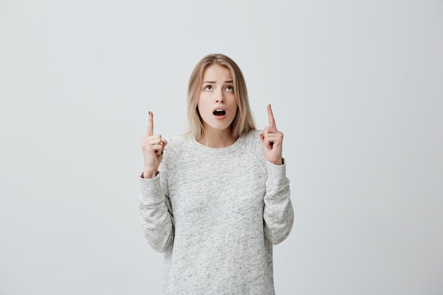 Astonished female with blonde dyed hair, in sweater looking and pointing upwards with her fingers being surprised to see something. Concerned excited young woman with wide opened mouth