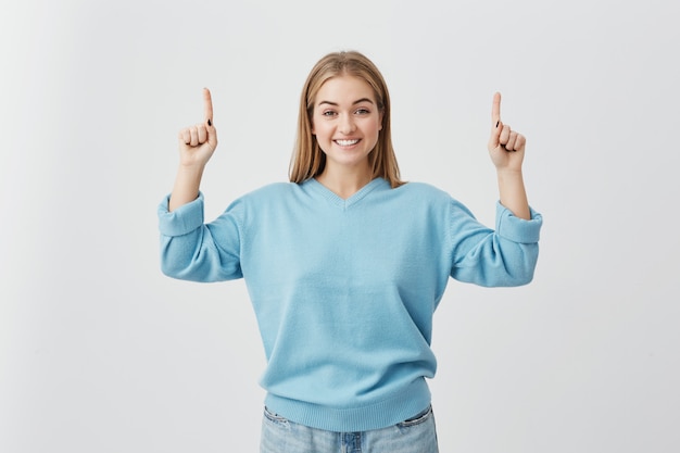 Astonished female with straight fair hair pointing upwards with her fingers being surprised to see something. Joyfuly smiling fair-haired woman posing .