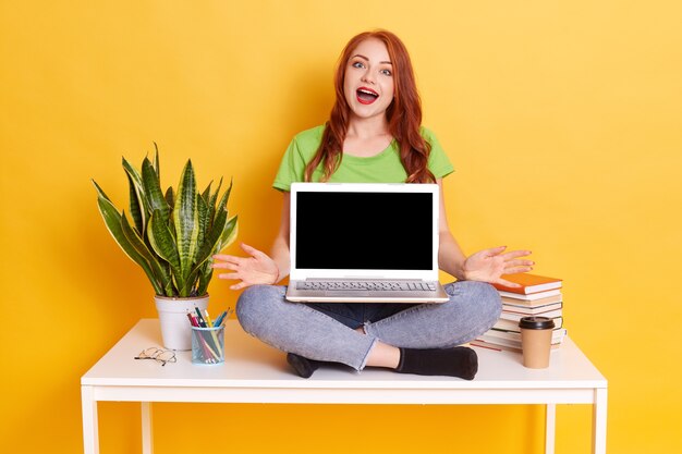 Удивленная женщина с широко открытым ртом, держащая ноутбук на коленях, показывая пустой экран, разводя руки в стороны, выглядит удивленной