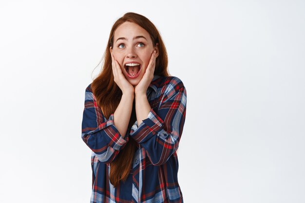 Astonished ginger girl screams from amazement and joy, looking at something awesome, look up and shouting happy, watching tv, standing on white