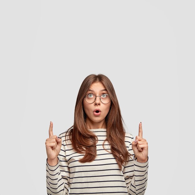 茶色の髪の驚いた印象的なヨーロッパの女性は、顎を落とし続け、2階を指して、広告コンテンツ用の空きスペースを示し、白い壁に立ち向かいます。広告