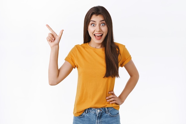 Astonished impressed fascinated young woman checking out cool promo opportunity of lifetime open mouth and smiling amused pointing upper left corner standing amazed white background