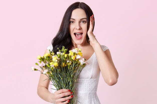 Astonished impressed young lady stands isolated over pink, opens mouth widely in surprise, holding bouquet of spring flowers