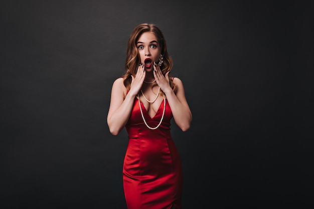 Astonished lady in red festive dress looks into camera Shocked woman in silk outfit posing on black isolated background