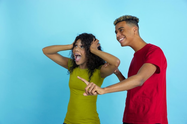 Astonished look at side like sport fans. Young emotional african-american man and woman in colorful clothes on blue background.