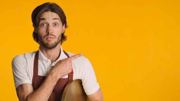 Astonished man wearing apron pointing aside with fingers isolated Young waiter looking amazed showing copy space over yellow background