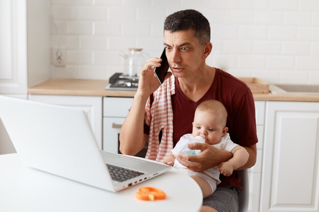 Astonished man wearing burgundy casual t shirt with towel on his shoulder, looks at laptop computer with big shocked eyes, looking after baby and working online from home.