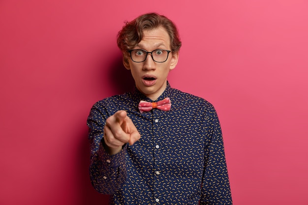 Astonished man with wavy hair, wears formal shirt and bowtie, points index finger directly , chooses someone, has startled expression, says hey you, looks with shock, isolated on pink