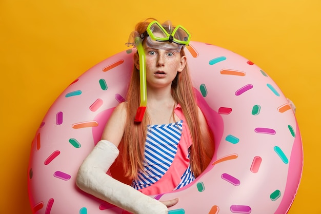 Astonished redhead freckled girl poses with inflated ring, wears swimsuit and snorkeling mask, shocked to see accident at sea, got trauma and injury during recreation, has broken arm in cast