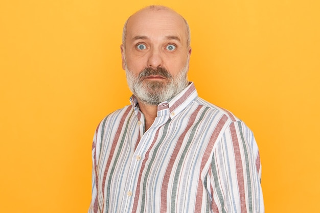 Free photo astonished senior bald unshaven man wearing striped shirt opening eyes wide being caught by surprise, receiving sudden unexpected news posing isolated against yellow studio wall background